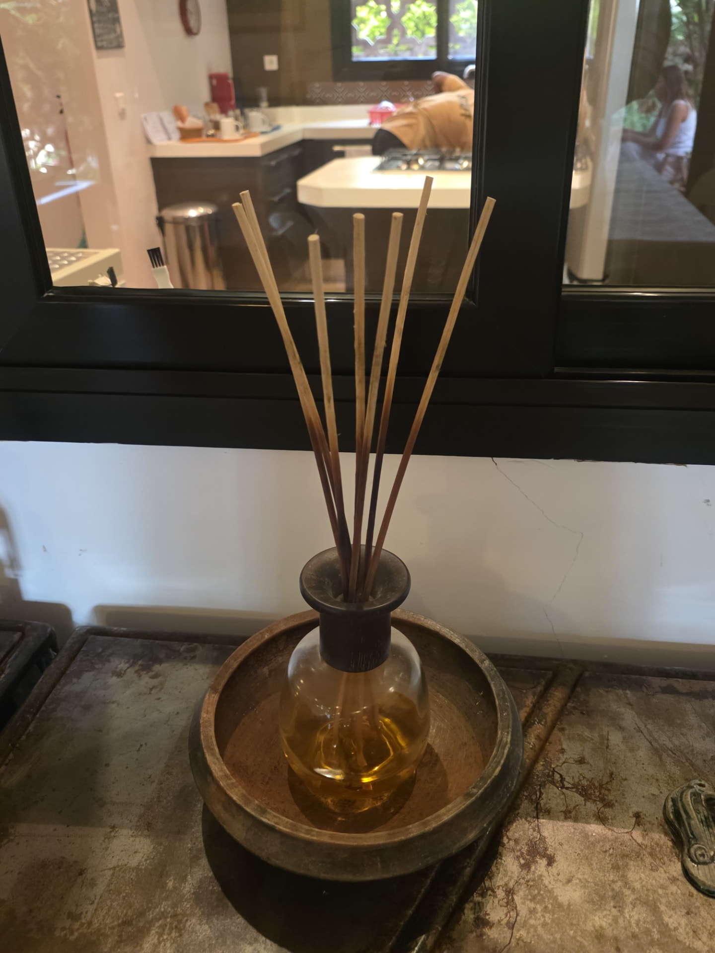 Flacon de parfum en verre avec de fines tiges sur un plateau rond devant une fenêtre intérieure