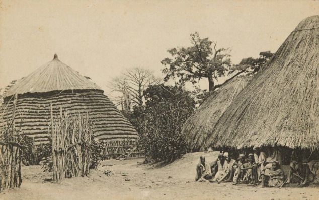 Carte postale d’archive légendée Timbo, Guinée, montrant une rue, des bâtiments en chaume et des personnes assises