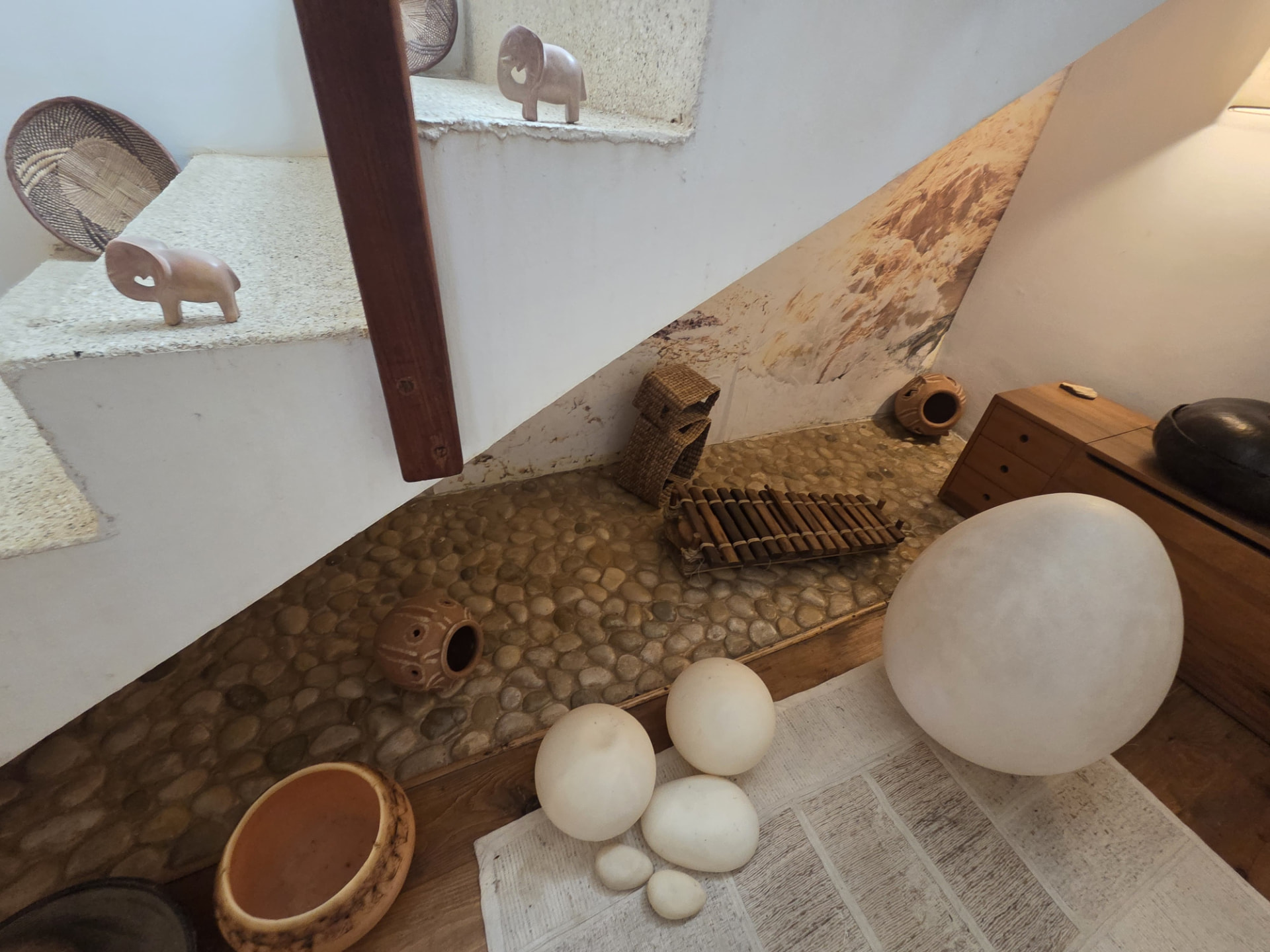 Présentation intérieure sous un escalier avec formes blanches arrondies, récipients en terre, animaux sculptés et instrument en bois