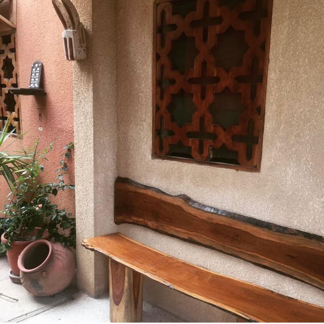 Banc en bois à bord naturel sous une fenêtre géométrique sculptée, avec plantes et jarres en terre