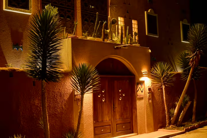 Carved wooden entrance doors of Maison Sahel lit warmly at night beside yucca palms