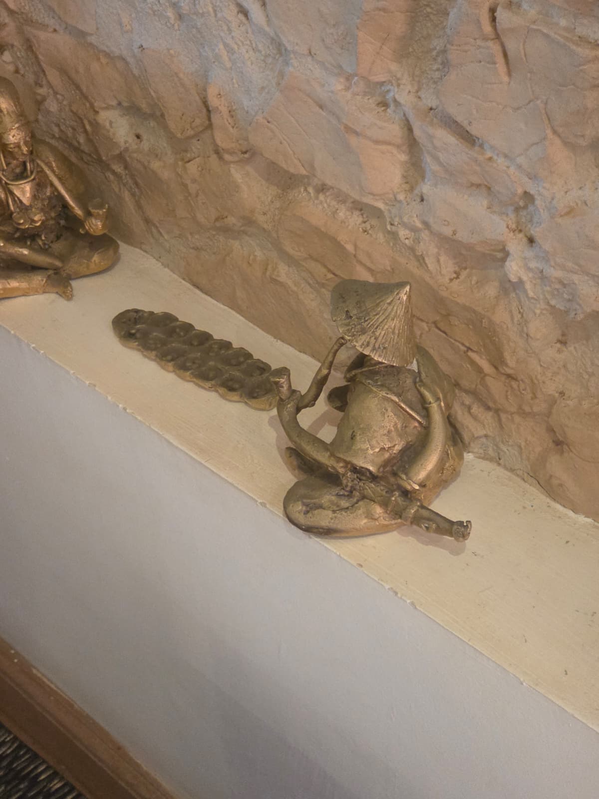 Petite figure en métal assise, coiffée d’un chapeau conique et tenant un instrument courbe sur un rebord